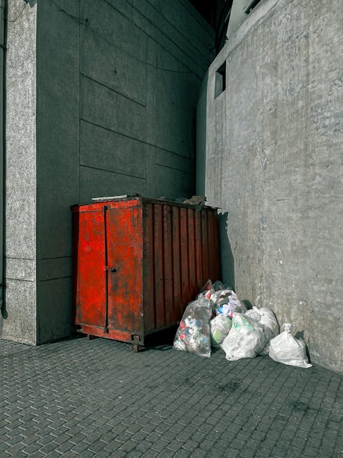 A red metal dumpster with visible rust spots and a slatted design is positioned against a dark green wall on a paved surface, with a light grey concrete wall on the right side. Several white plastic rubbish bags filled with waste are piled next to the dumpster, some leaning against its side. The surrounding environment suggests an urban alleyway or back street, with minimal natural light and no visible signage. The scene relates to private waste collection and the challenges of managing bulky or excess refuse outside scheduled rubbish removal services, exemplifying the need for alternative waste handling methods like independent disposal or on-site clearance. The overall atmosphere is utilitarian, focusing on the practical aspect of rubbish storage and disposal within a city environment, fitting with the topic of avoiding hidden costs associated with Wimbledon rubbish clearance services. Waste Disposal Wimbledon occasionally handles this type of waste, providing flexible solutions for such scenarios.