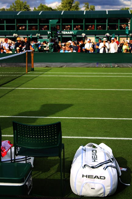 A view of a grass tennis court with clearly marked white boundary lines, partially shaded by shadows cast from the surrounding environment. In the foreground, a dark green plastic chair with a perforated backrest is positioned next to a small green container and a backpack with the brand name 'HEAD' printed on it, resting on the grass near the net. To the left side of the image, a red and white sports water bottle is visible beside the chair. In the background, beyond the court, there is a green seating area or viewing platform filled with spectators sitting and standing, some wearing hats and casual clothing. The seating structure is elevated, multi-tiered, and constructed with metal and green panels, with the word 'TENAR' partially visible on a sign attached to it. The overall scene suggests a sports event or tennis match, and while primarily focused on the court and seating area, this setting could relate to external maintenance or temporary storage related to sports or event organization, consistent with the context of waste management or private event rubbish handling by companies such as Waste Disposal Wimbledon.