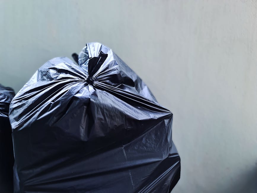 Close-up image of a tightly tied black plastic rubbish bag placed against a plain, light-colored wall. The bag appears to be made of glossy plastic material, with visible creases and folds, and is filled with waste, giving it a slightly rounded, bulging shape. In the background, portions of additional black rubbish bags are partially visible, stacked or placed nearby, indicating a collection or disposal scene. The environment looks like an indoor or sheltered area, possibly a storage corner or utility space, where waste is gathered before collection. The lighting is even, without strong shadows, highlighting the texture of the plastic and the smooth, matte finish of the wall behind. This scene aligns with practices in independent rubbish disposal, handled by waste management services like Waste Disposal Wimbledon, where private collection or on-site clearance may be arranged outside of local authority services.