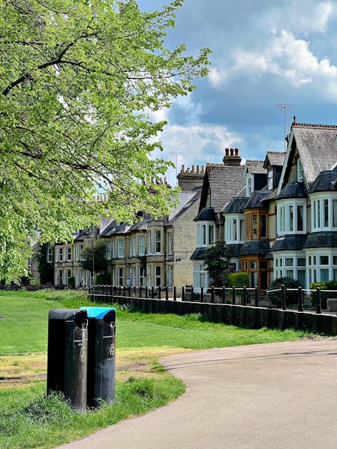 A pair of black public rubbish bins with blue and green lids are positioned on a grassy verge next to a paved footpath in a residential area. Behind the bins, there's a row of Victorian-style terraced houses with bay windows, brick facades, and slate roofs. The houses are closely spaced, with small front gardens enclosed by low wooden fences, some featuring shrubs and small trees. A large, leafy tree with fresh green foliage extends its branches across the upper part of the image, providing partial shade. The sky above is partly cloudy with patches of blue visible between white clouds. The scene appears calm and tidy, characteristic of an urban residential suburb, with no visible waste or rubbish in the vicinity, aligning with private waste collection services that may handle rubbish removal for the neighborhood. Waste Disposal Wimbledon might assist with on-site clearance or private waste collection needs for similar properties, emphasizing professional rubbish management. The peaceful environment underscores the importance of proper waste disposal infrastructure in residential areas. The image conveys a neat, organized streetscape suitable for a location served by independent rubbish collection services, contributing to neighborhood cleanliness and proper waste handling.